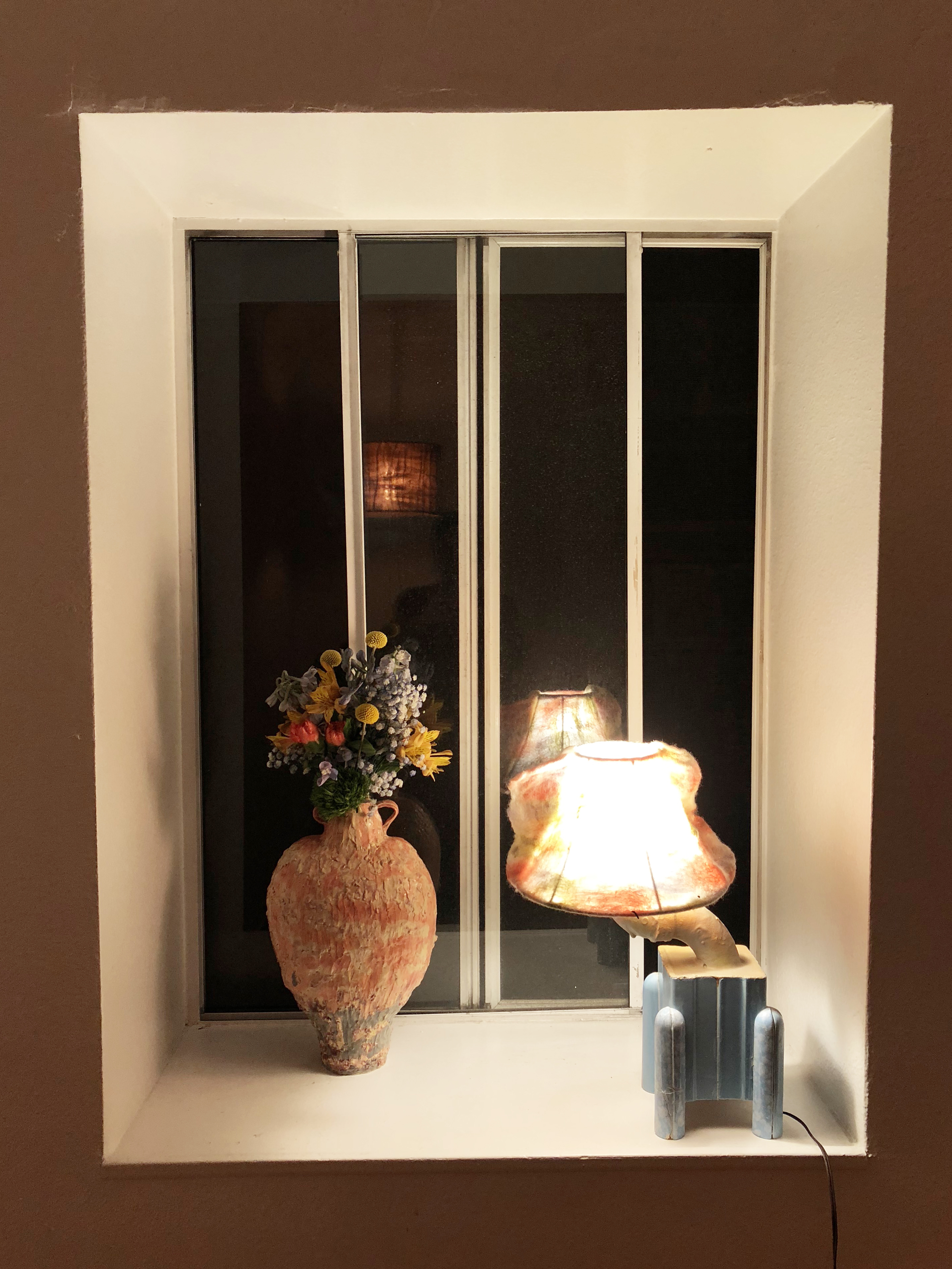 An orange ceramic vase with flowers and a felt lamp on a windowsill